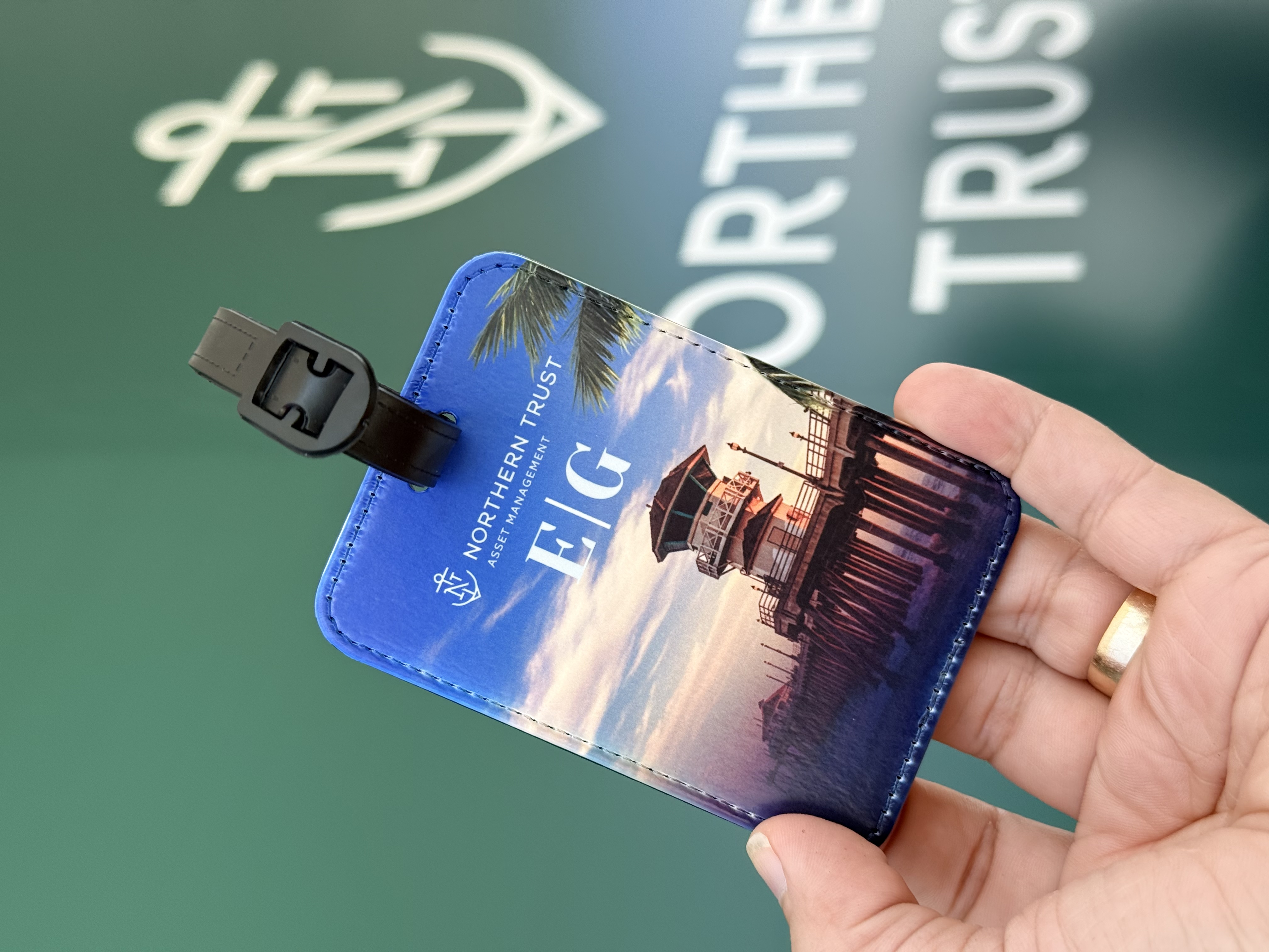 A luggage tag monogrammed E G held in front of the Northern Trust Asset Management booth wall